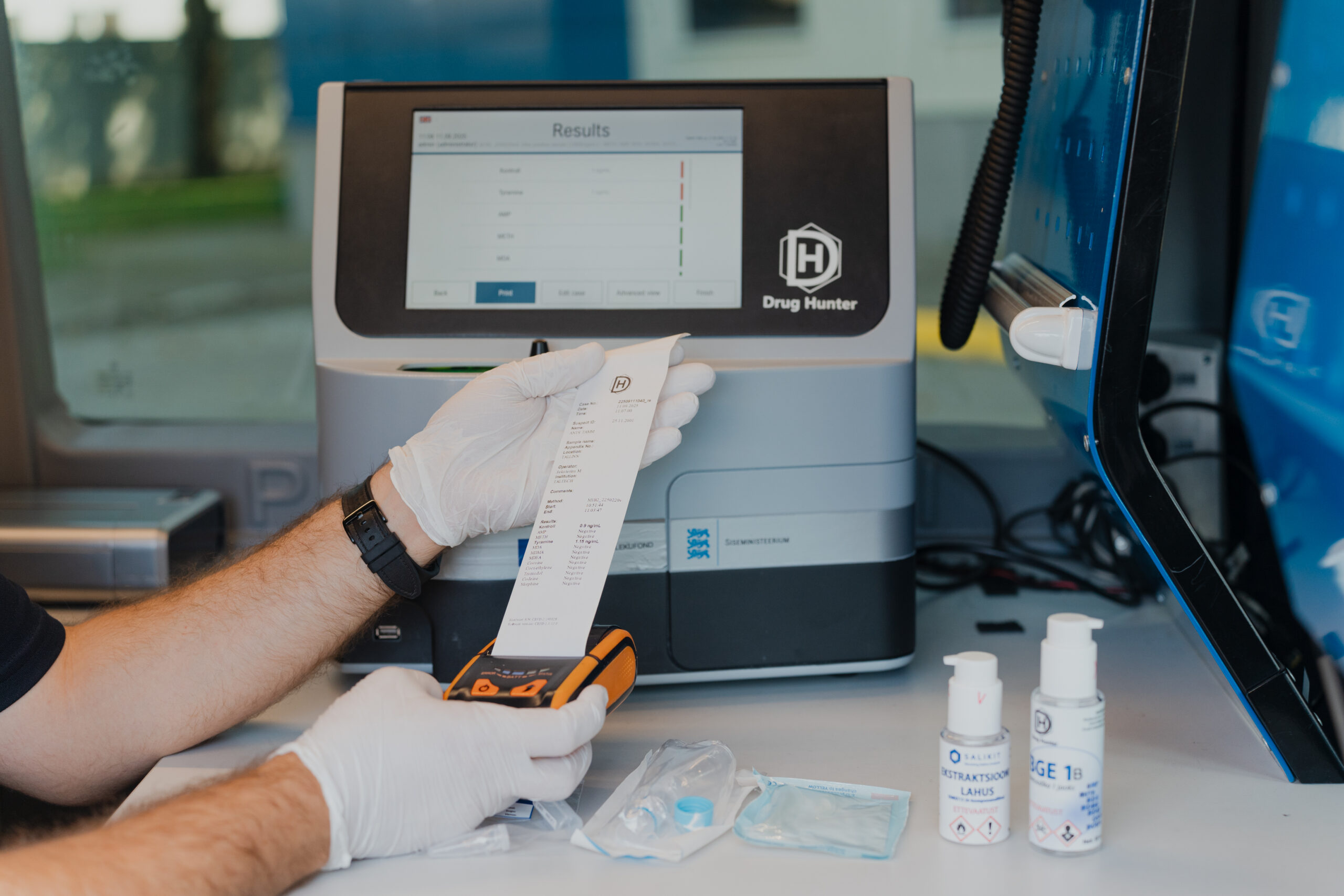 Gloved operator holds a printed test result next to the Drug Hunter analyzer and reagent bottles
